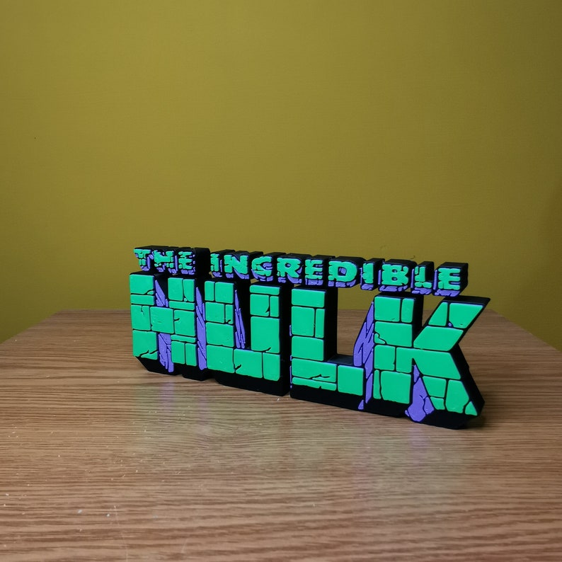 K&ouml;nnte beinhalten: Ein 3D-gedrucktes Modell des "The Incredible Hulk"-Logos. Das Logo besteht aus gr&uuml;nem und violettem Kunststoff und hat eine Ziegelstruktur. Das Logo steht auf einer Holzoberfl&auml;che.