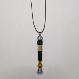 May include: A silver, black, and gold lightsaber pendant necklace. The pendant has a detailed design with a black grip, gold accents, and a yellow stripe. The necklace is suspended from a black cord.