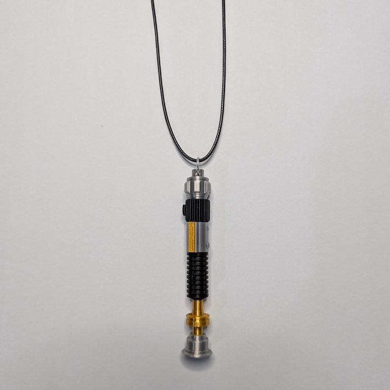 May include: A silver, black, and gold lightsaber pendant necklace. The pendant has a detailed design with a black grip, gold accents, and a yellow stripe. The necklace is suspended from a black cord.