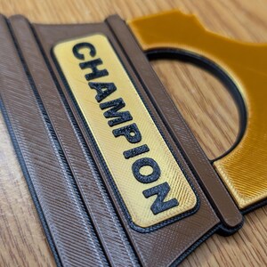 May include: Close-up of a brown and gold trophy with the word "CHAMPION" printed on the front.