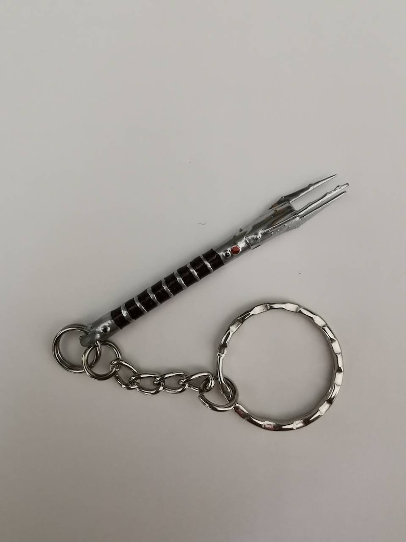 May include: A silver and black lightsaber keychain with a red gem. The keychain has a silver chain and a silver ring.