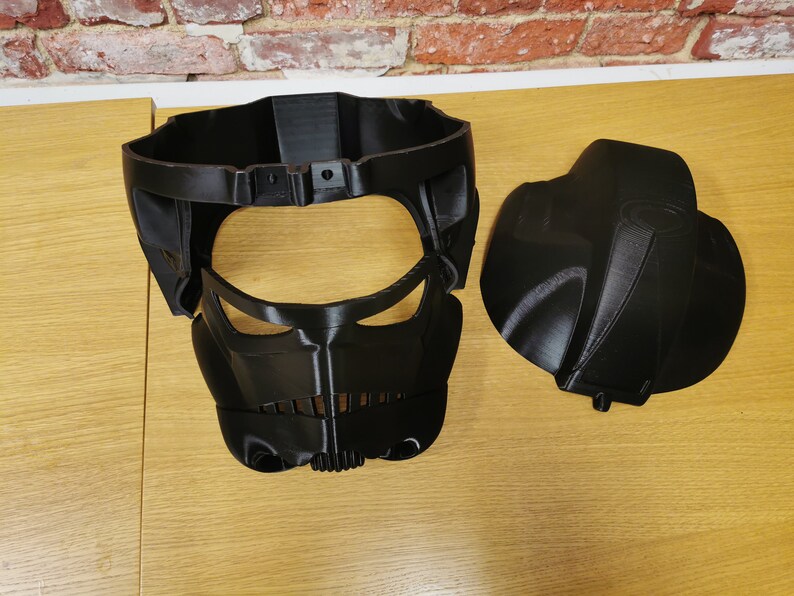 May include: Two black plastic pieces of a 3D printed helmet. The helmet has a face mask with a visor and a separate top piece.