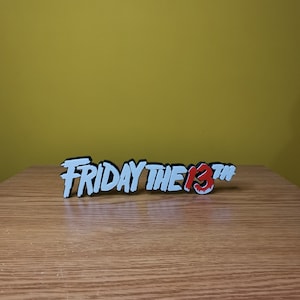 Puede incluir: Un letrero impreso en 3D en blanco y negro que dice "FRIDAY THE 13TH" con un "13" rojo sobre una superficie de madera.