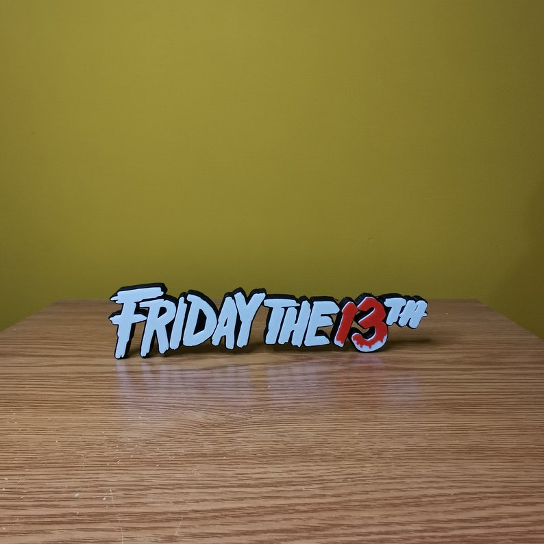 Puede incluir: Un letrero impreso en 3D en blanco y negro que dice "FRIDAY THE 13TH" con un "13" rojo sobre una superficie de madera.