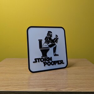 May include: A white square sign with a black border. The sign features a black and white illustration of a Stormtrooper sitting on a toilet. The text "Storm Pooper" is printed in black below the illustration.
