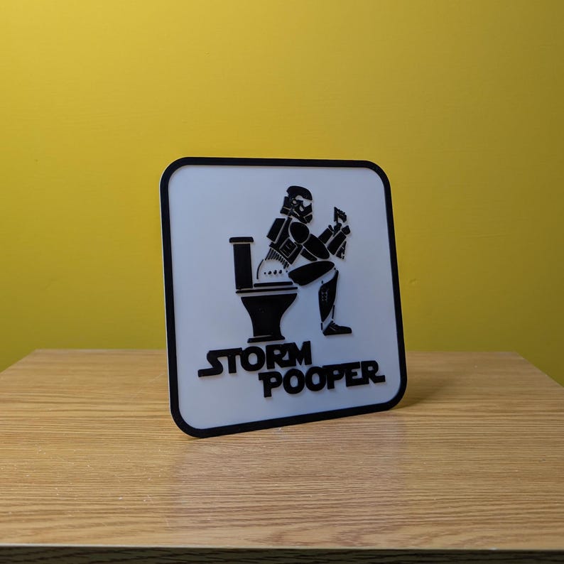 May include: A white square sign with a black border. The sign features a black and white illustration of a Stormtrooper sitting on a toilet. The text "Storm Pooper" is printed in black below the illustration.