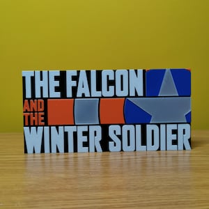 May include: A black and white sign with the text "The Falcon and the Winter Soldier" in white letters. The sign has a blue, white, and red star design in the upper right corner.