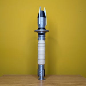 May include: A white and silver lightsaber prop with a black handle and a silver tip. The lightsaber is standing upright on a wooden surface.