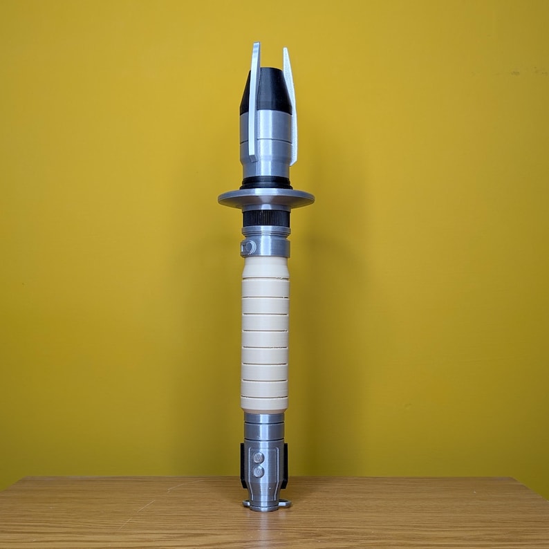 May include: A white and silver lightsaber prop with a black handle and a silver tip. The lightsaber is standing upright on a wooden surface.