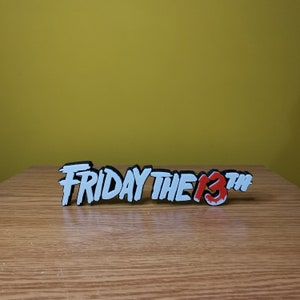 Puede incluir: Un letrero impreso en 3D blanco y rojo que dice "FRIDAY THE 13TH" en una fuente estilizada. El letrero est&aacute; sobre una superficie de madera.
