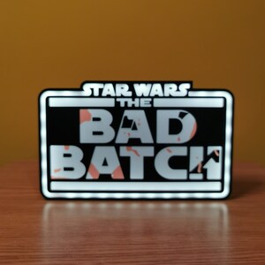 May include: A black and white sign with white LED lights around the border. The sign reads "Star Wars The Bad Batch".