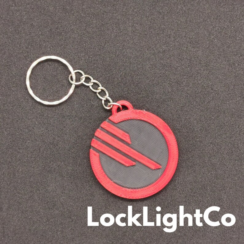 May include: A red and black keychain featuring the Rebel Alliance symbol from Star Wars. The keychain is made of plastic and has a silver metal ring.