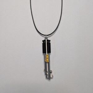 May include: A silver-colored lightsaber pendant with black and gold accents hangs from a black cord. The pendant is a detailed miniature replica of a lightsaber, a popular item in science fiction.