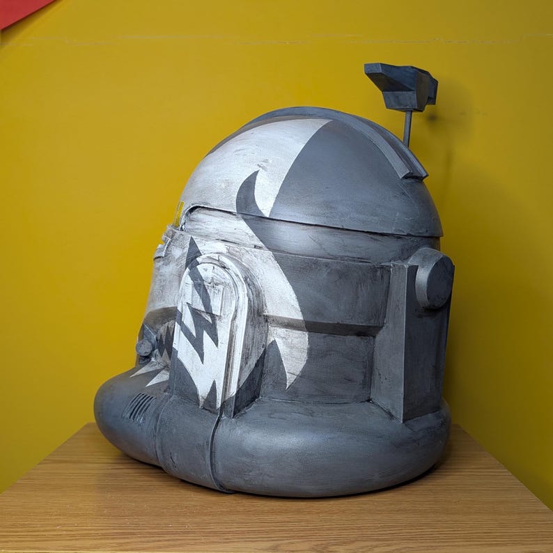 May include: A gray and white Star Wars clone trooper helmet with a black and white pattern. The helmet has a small antenna on top.