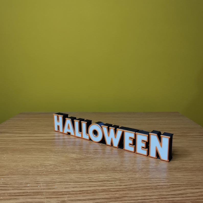 K&ouml;nnte beinhalten: Ein schwarzes und wei&szlig;es "HALLOWEEN"-Schild mit orangefarbenem Rand. Das Schild ist aus Kunststoff und steht auf einer Holzoberfl&auml;che.