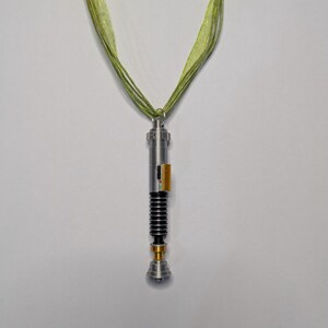 May include: A silver-colored lightsaber pendant with black and gold accents, suspended from a green ribbon necklace. The lightsaber features a detailed design with a ribbed handle and a gold-colored base.