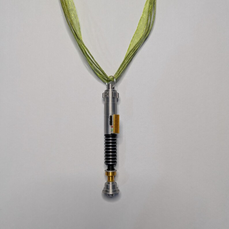 May include: A silver-colored lightsaber pendant with black and gold accents, suspended from a green ribbon necklace. The lightsaber features a detailed design with a ribbed handle and a gold-colored base.