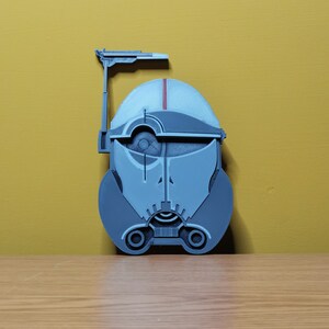 Pu&ograve; includere: Un modello stampato in 3D di un elmetto di un clone trooper di Star Wars. L'elmetto &egrave; grigio e ha una striscia rossa sulla parte superiore. L'elmetto ha una visiera e una piccola antenna sulla parte superiore.