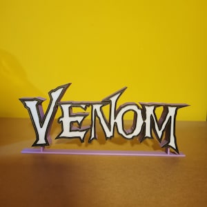 May include: A 3D-printed sign spelling "VENOM". The letters are white with black outlines, set on a light purple base. The background is a solid yellow. The sign is designed in a stylized font.