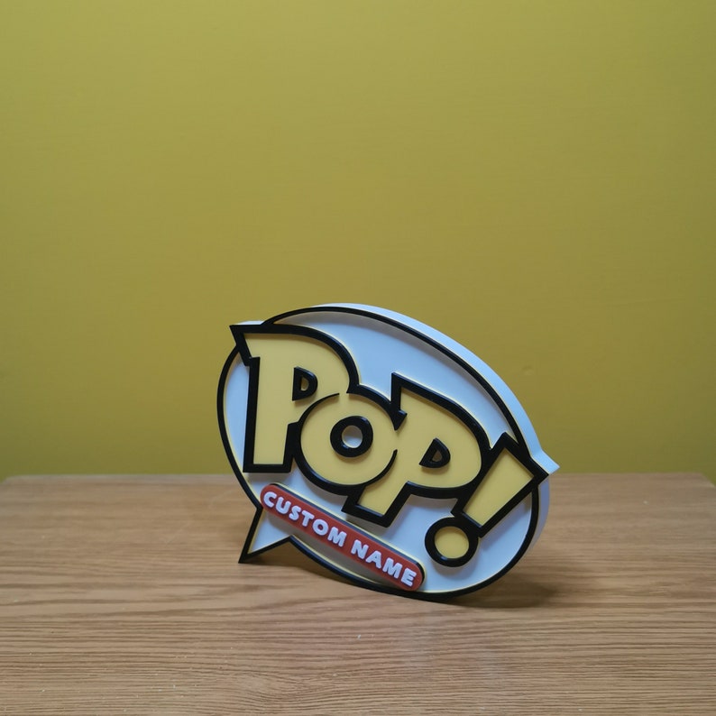 May include: A white and yellow 3D printed Pop! figure with black lettering. The figure is shaped like a speech bubble with the word "Pop!" in black lettering and the words "Custom Name" in red lettering.