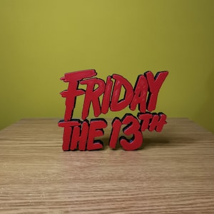 May include: Red 3D printed sign that reads "Friday the 13th" in a stylized font. The sign is on a wooden surface.