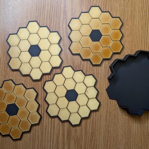 May include: Four black and gold honeycomb-shaped coasters with a black center circle. The coasters are arranged in a semi-circle on a wooden surface. A black hexagonal storage box is also shown.