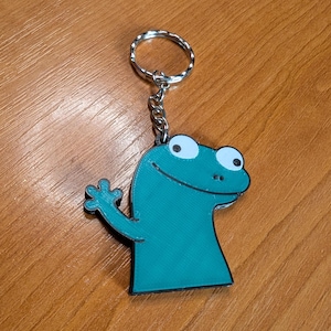 Pode incluir: Chaveiro de sapo de desenho animado azul-petróleo, com argola e corrente prateadas. O sapo tem olhos brancos e contorno preto. O sapo está acenando com a mão direita. Sobre uma superfície de madeira.
