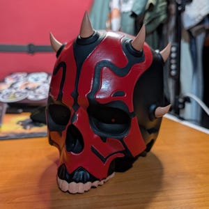 May include: A red and black skull mask with curved horns. The mask has a detailed design with red markings on a black base, and tan-colored horns. The mask is likely a costume or collectible item.