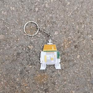 Puede incluir: Un llavero de robot blanco y amarillo con una cadena plateada. El robot tiene un dise&ntilde;o verde y amarillo en la cabeza y el cuerpo.