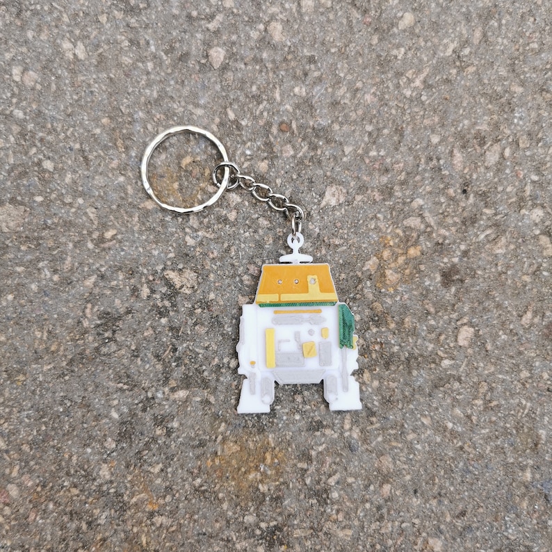 Puede incluir: Un llavero de robot blanco y amarillo con una cadena plateada. El robot tiene un dise&ntilde;o verde y amarillo en la cabeza y el cuerpo.