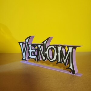 May include: A 3D-printed sign spelling "Venom" in a stylized font. The white letters have black outlines, set against a light purple background. The sign is on a light purple base, with a yellow background.