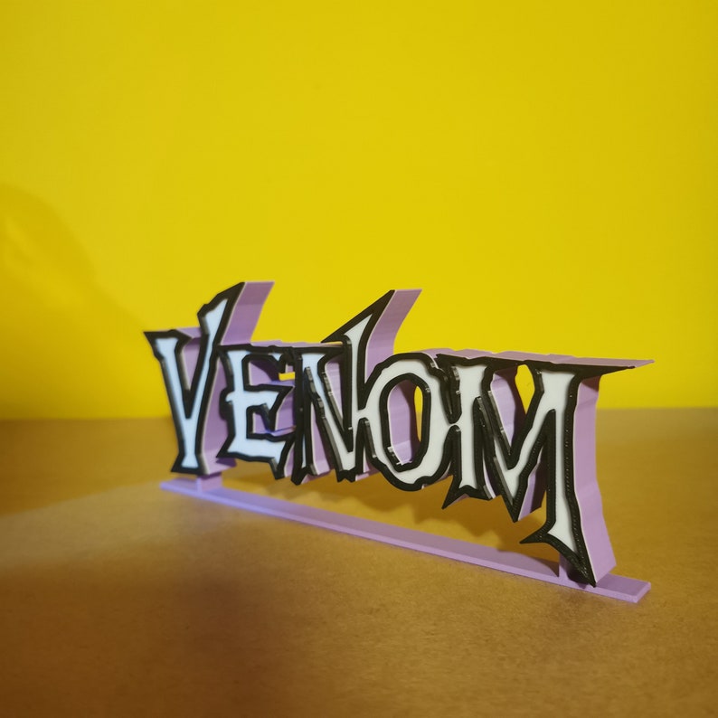 May include: A 3D-printed sign spelling "Venom" in a stylized font. The white letters have black outlines, set against a light purple background. The sign is on a light purple base, with a yellow background.