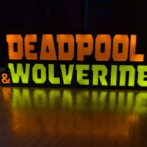 Pu&ograve; includere: Un cartello rettangolare nero con le parole "DEADPOOL & WOLVERINE" in lettere illuminate arancioni e verdi. Il cartello &egrave; su una superficie di legno, con le parole riflesse.