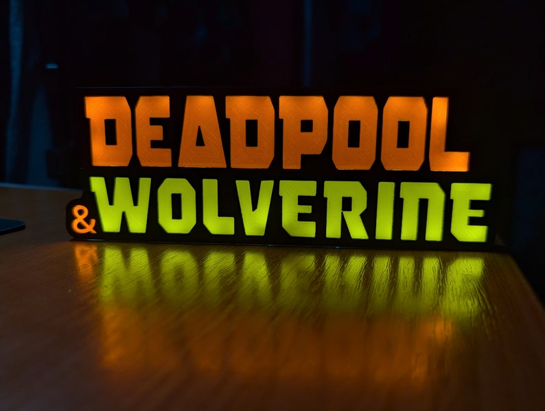 Pu&ograve; includere: Un cartello rettangolare nero con le parole "DEADPOOL & WOLVERINE" in lettere illuminate arancioni e verdi. Il cartello &egrave; su una superficie di legno, con le parole riflesse.