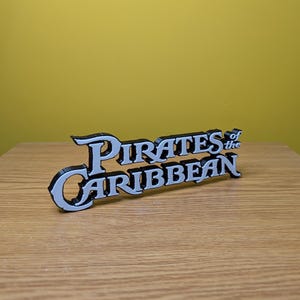 May include: A 3D printed sign that reads "Pirates of the Caribbean" in white letters with black outlines.