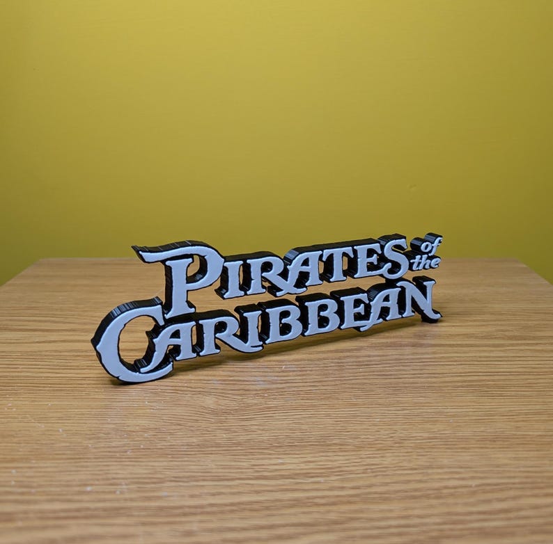 May include: A 3D printed sign that reads "Pirates of the Caribbean" in white letters with black outlines.