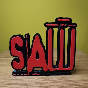 May include: Black and red 3D wall decor with the word "SAW" in bold letters. The letters are outlined in black and filled in with red. The "S" and "A" are in a stylized font.
