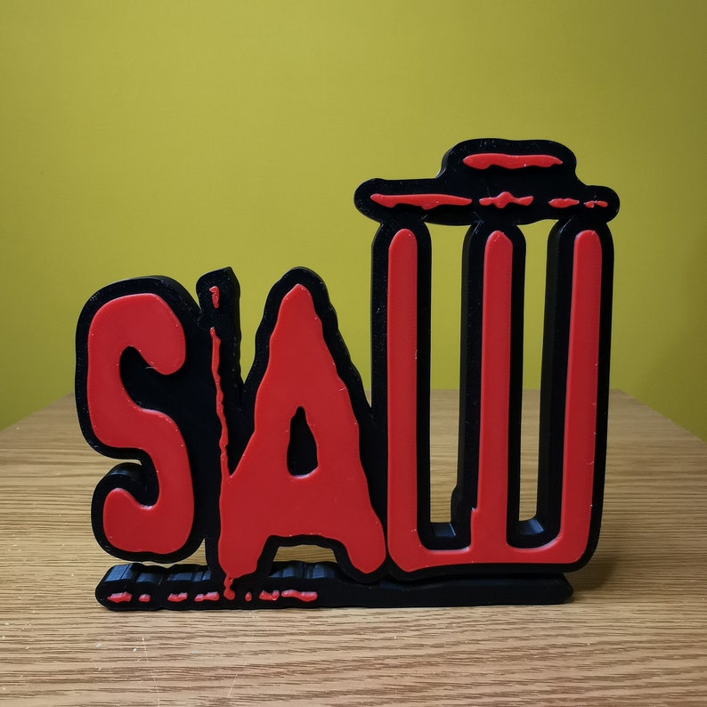 May include: Black and red 3D wall decor with the word "SAW" in bold letters. The letters are outlined in black and filled in with red. The "S" and "A" are in a stylized font.