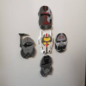May include: Five Star Wars clone trooper helmet magnets in gray, white, red, and black. The helmets are all different designs and are attached to a white surface.