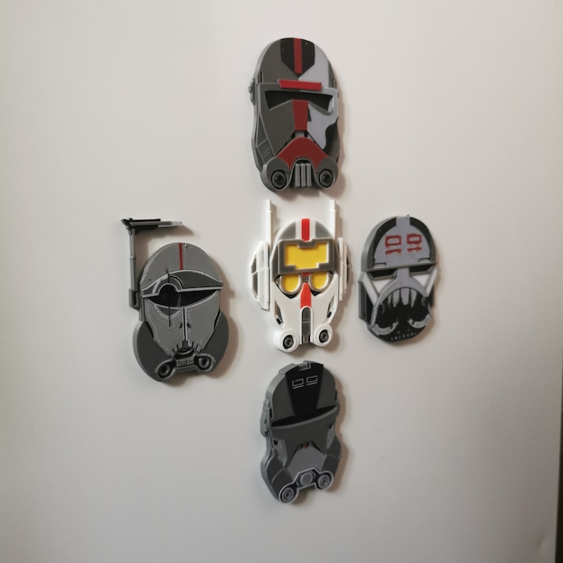 May include: Five Star Wars clone trooper helmet magnets in gray, white, red, and black. The helmets are all different designs and are attached to a white surface.