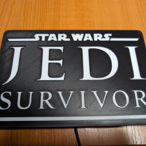 以下が含まれることがあります： 「STAR WARS JEDI SURVIVOR」と白文字で書かれた黒い長方形の看板です。