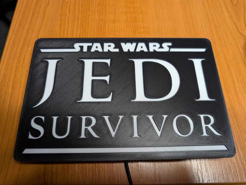 以下が含まれることがあります： 「STAR WARS JEDI SURVIVOR」と白文字で書かれた黒い長方形の看板です。