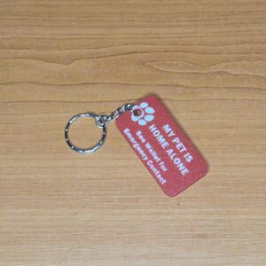 May include: Red rectangular keychain with white text that reads "My Pet is Home Alone" and "See Wallet for Emergency Contact". A paw print symbol is above the text.
