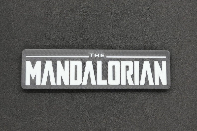 May include: A black and white rectangular patch with the text "THE MANDALORIAN" in white letters.