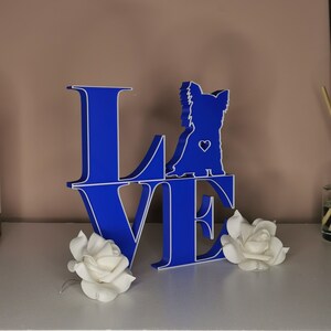 May include: Blue wooden "LOVE" sign with a white outline. A silhouette of a dog is standing on the "O". The sign is on a white surface with two white roses.