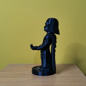 May include: A black plastic figurine of Darth Vader from Star Wars. The figurine is standing on a black base and has a cape.