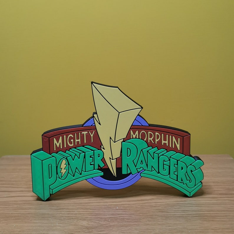 Pu&ograve; includere: Un logo colorato dei Power Rangers con la scritta "Mighty Morphin Power Rangers". Il logo presenta un fulmine giallo, testo verde e rosso e uno sfondo blu e nero. Il logo &egrave; 3D e si trova su una superficie di legno.