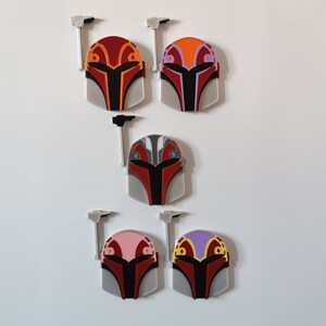 May include: Five Mandalorian helmet pins with different colored accents. The helmets are red, orange, gray, pink, and purple. Each helmet has a silver antenna.
