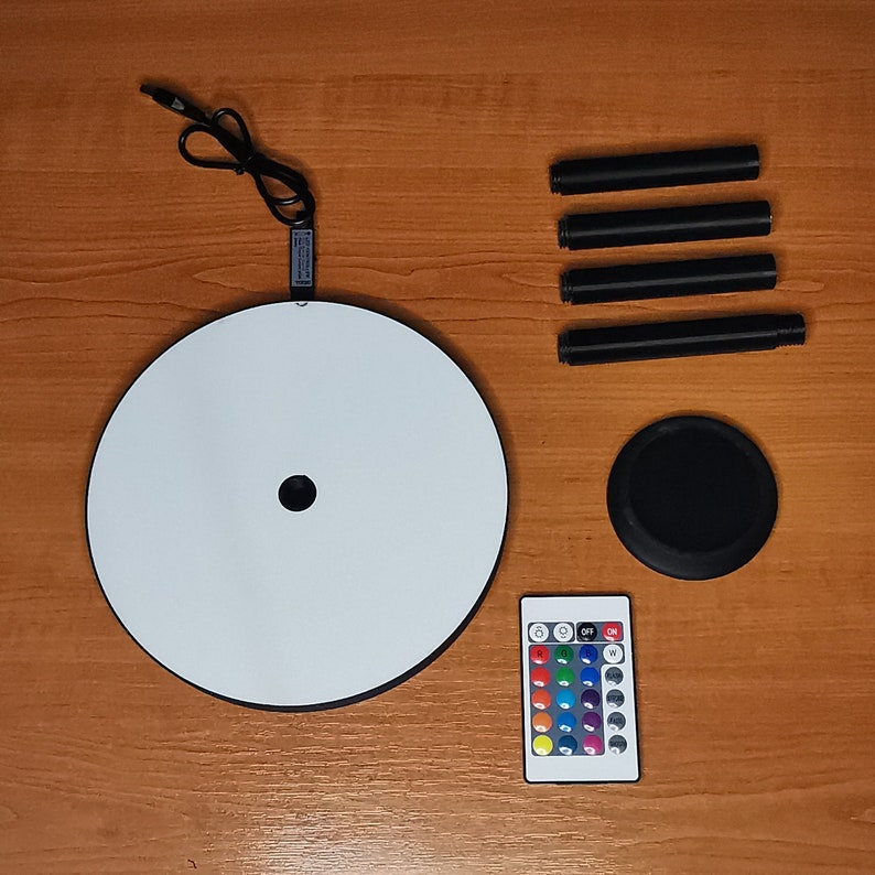 May include: A white round light base with a black remote control with color changing buttons. The light base is connected to a black power cord and has four black metal legs.