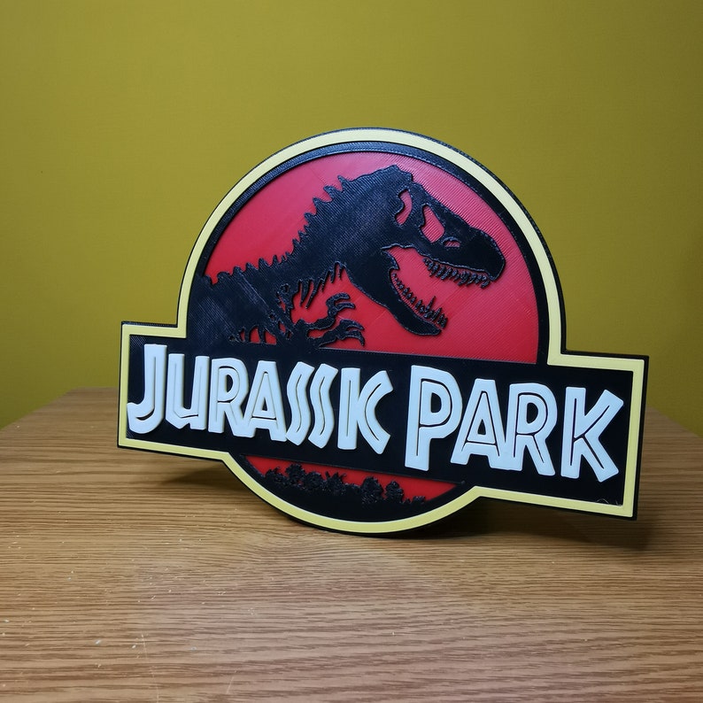 May include: A 3D printed Jurassic Park logo with a black dinosaur silhouette on a red background. The logo is surrounded by a yellow border and the words "Jurassic Park" are printed in white.